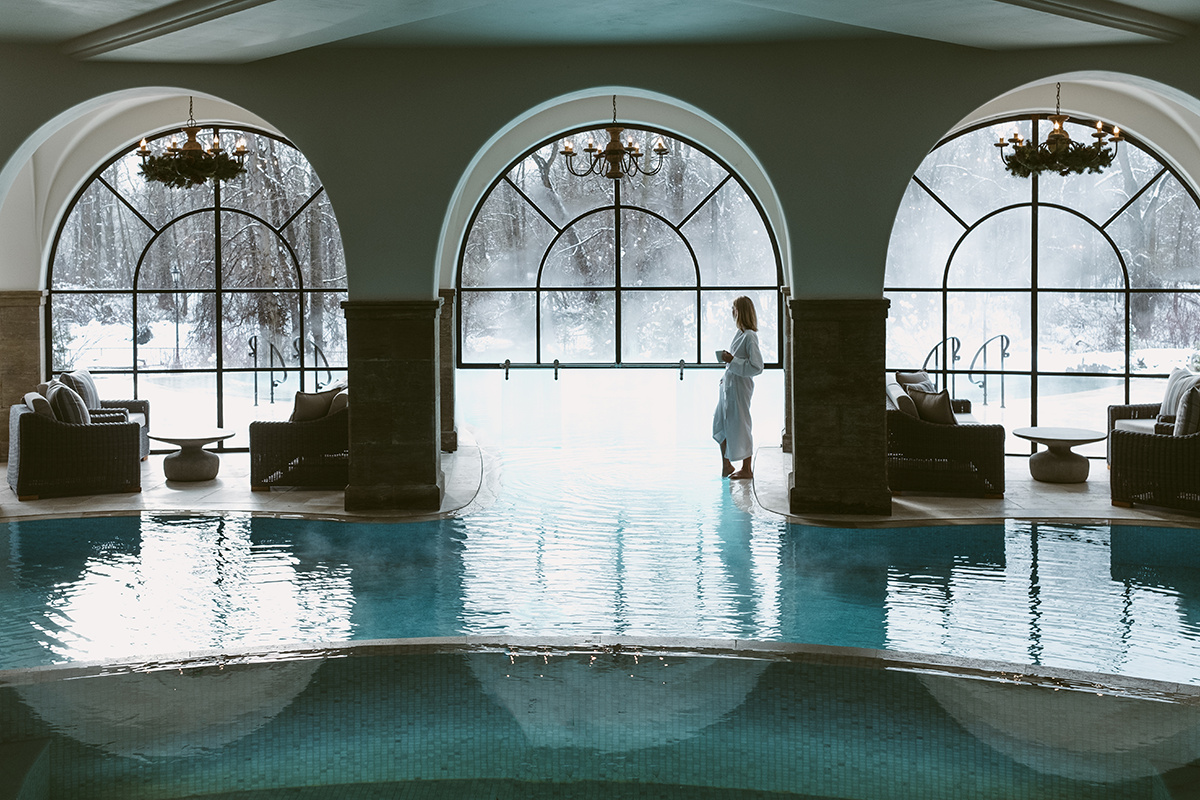 A large indoor pool with arched windows and chandeliers, a seating area, and a snowy view outside.