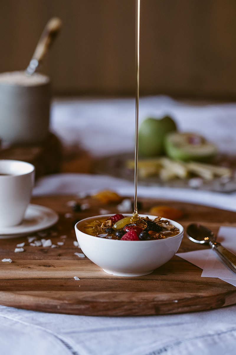 A bowl of oatmeal with fruit and honey being poured over it on a wooden board