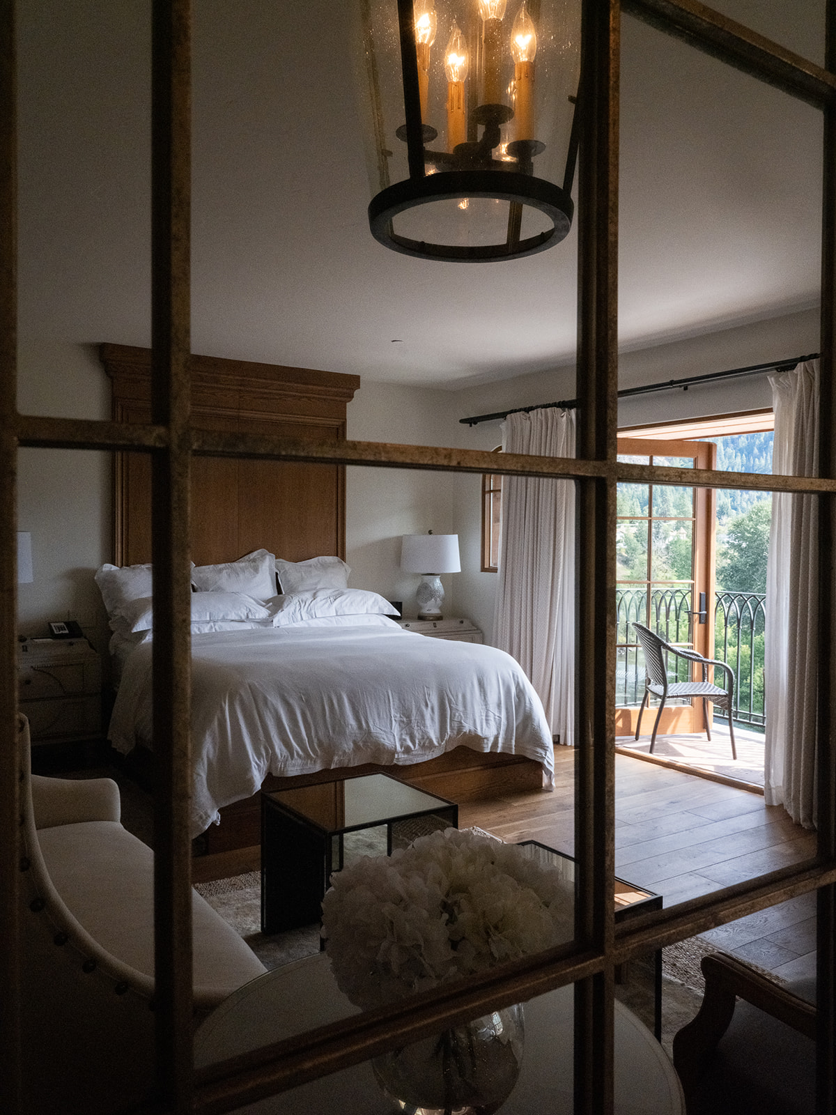 A hotel room with a large bed, chandelier, balcony, sofa, and table.