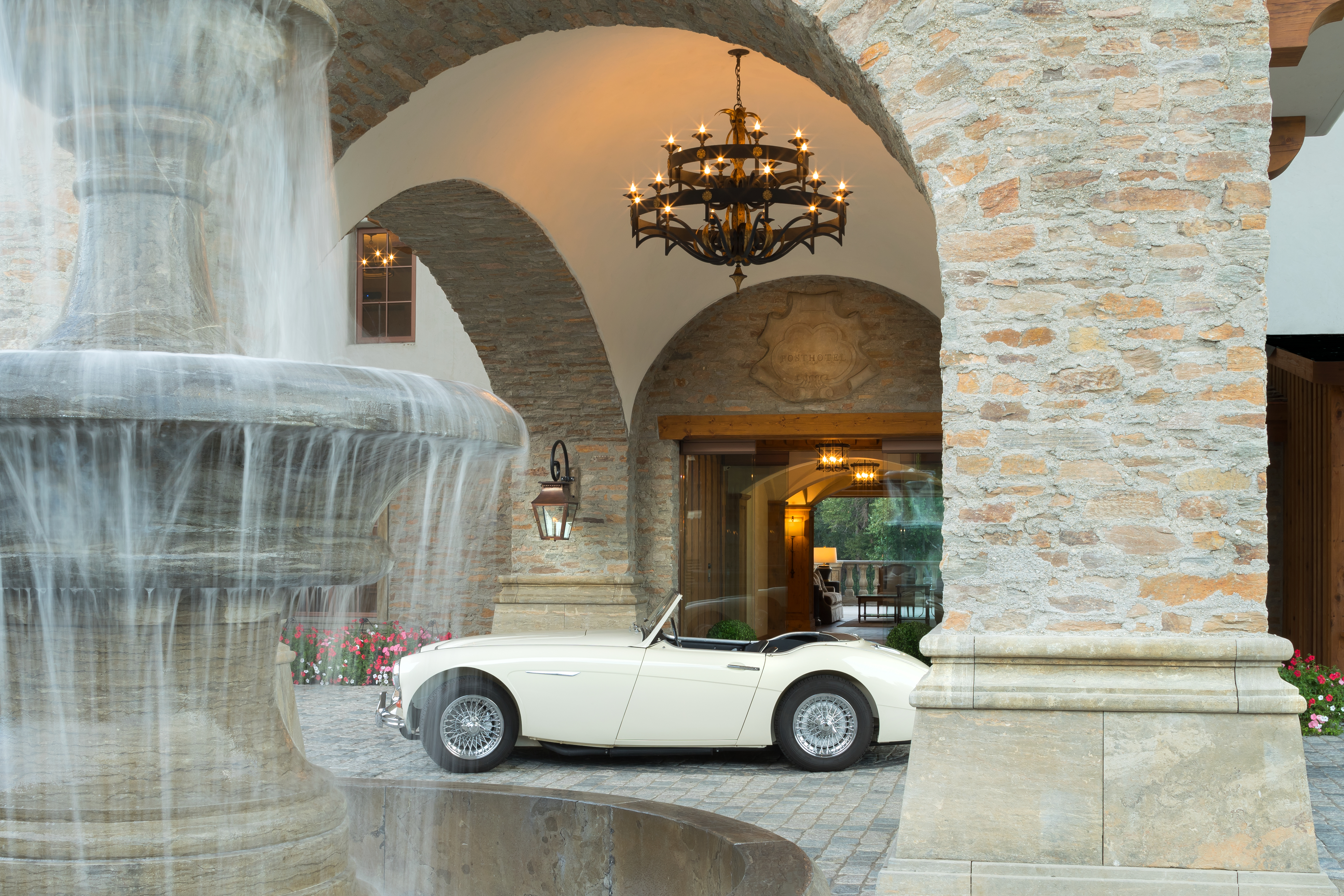 A white convertible car parked in front of a stone building with a fountain and chandelier.