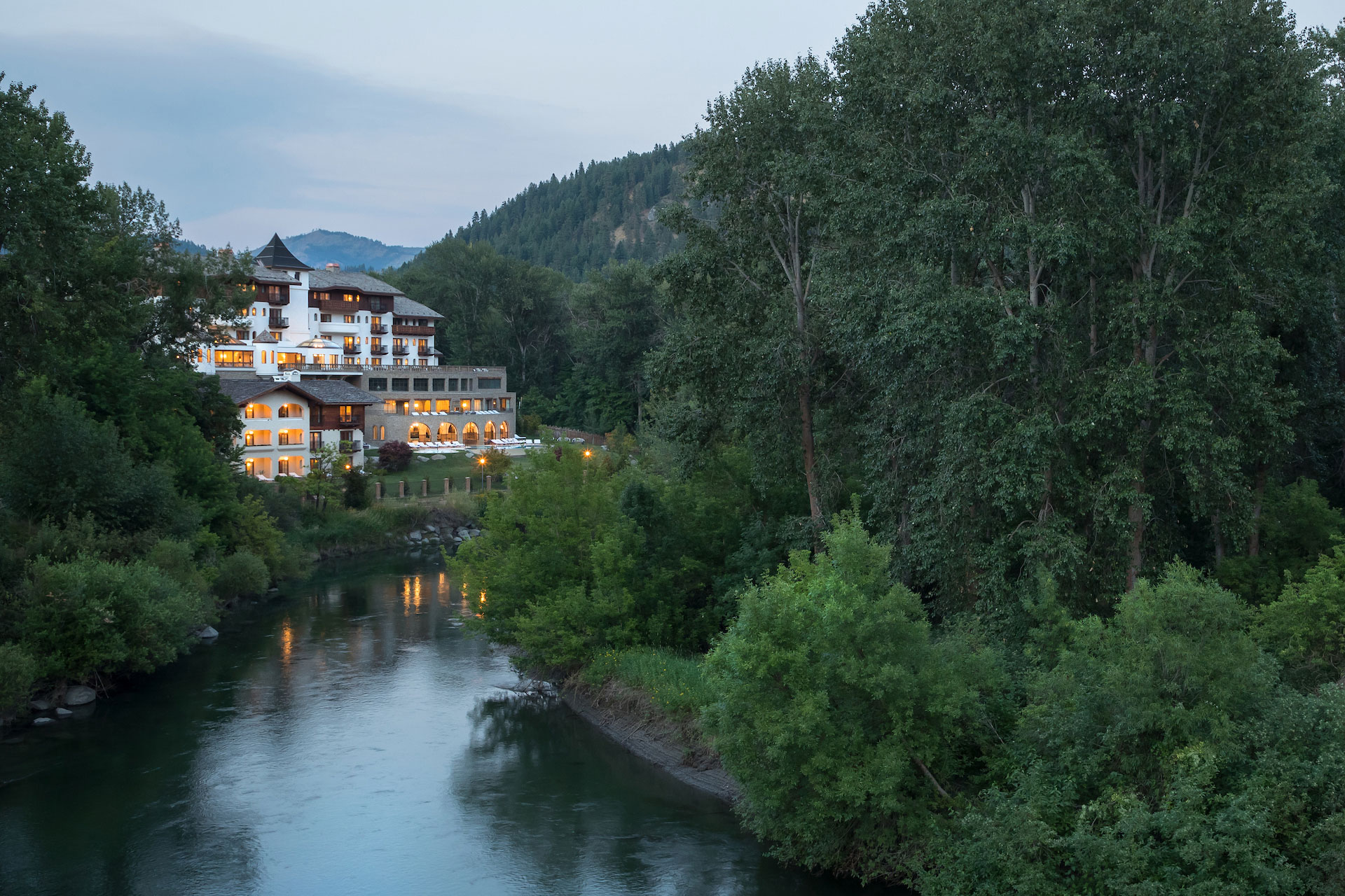 A large building with many windows sits on the riverbank, surrounded by trees and mountains.